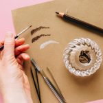 Close-up of a hand holding a brush with makeup tools on colored paper.